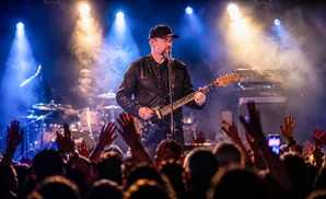 A man in a black baseball cap plays an electric guitar and sings into a microphone on a smoky stage with bright lights.