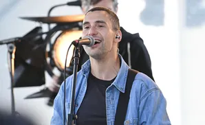 A man in a denim jacket sings into a microphone on stage with a band.