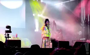 A woman in a puffy, colorful jacket sings into a microphone on a stage, with lights and bubbles in the background.