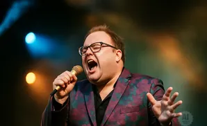 A man in a floral jacket sings into a microphone with his mouth open.