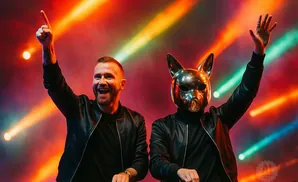 Two DJs, one with a metal dog mask, perform under colorful stage lights.