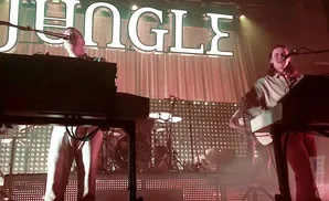 Two men playing keyboards on stage in front of a backdrop with the word 