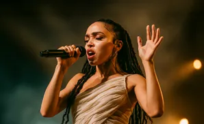Singer FKA twigs performs on stage, holding a microphone and gesturing with her other hand.
