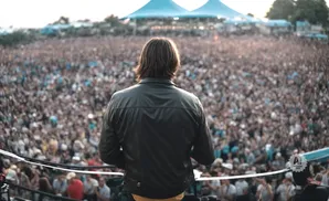 A person in a black leather jacket stands with their back to the camera, facing a large crowd at an outdoor concert.