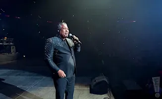 A man in a sparkly black suit sings into a microphone on a stage with a blurred audience in the background.