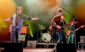 A band plays on stage, with a singer in the foreground holding a guitar and extending his arms.