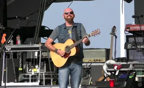 A bald man with a beard and sunglasses plays an acoustic guitar on stage.