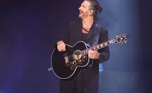 A smiling man with a bun in his hair plays an acoustic guitar on stage.