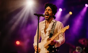 A male singer in a ruffled white shirt and black turban plays a white electric guitar on stage under purple spotlights.