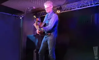 Man with gray hair and glasses playing an acoustic guitar on stage.
