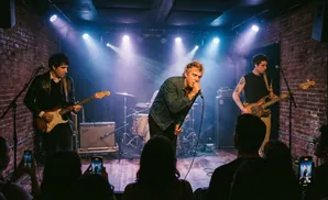 A band performs on a brick stage with spotlights, a singer in the center, a guitarist to the left, and a bassist to the right.
