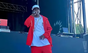 Rapper in red hoodie and pants performs on stage, wearing a white hat and gold chain.