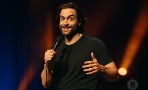 A comedian on stage holds a microphone and gestures with his hands while wearing a black t-shirt.