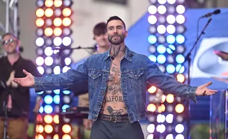 Singer Adam Levine performs shirtless in a denim jacket, his tattooed torso visible, with a backdrop of colorful lights.