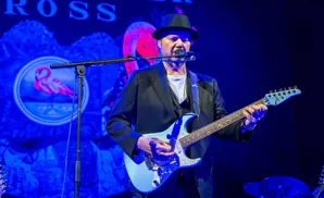 Musician in a hat and suit plays a blue guitar on stage with a flamingo logo backdrop.