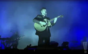 A man in a suit plays an acoustic guitar on a stage with blue lighting and smoke.