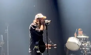 A man with a beard and glasses plays a harmonica into a microphone while a drummer plays in the background.