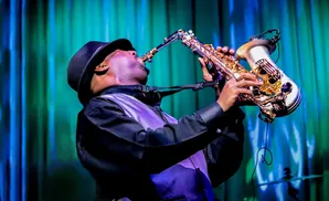 A man in a black hat and vest plays a saxophone on a stage with a blue and green backdrop.