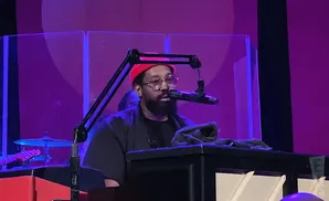 A Black man wearing a red beanie and glasses plays a keyboard, with a microphone in front of him.