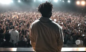 Man with dreadlocks on stage facing a large, cheering crowd with hands raised.