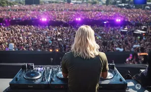 A DJ with long blonde hair plays music for a massive crowd at an outdoor festival.
