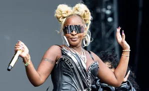 Mary J. Blige in a black leather outfit, sunglasses, and blonde hair in buns, holding a gold microphone.