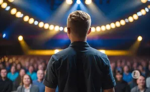 A man in a dark shirt stands with his back to the camera, facing a blurred audience under a stage with yellow lights.