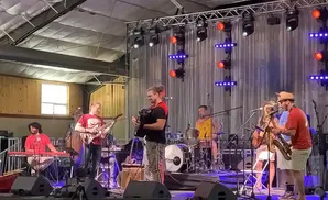 A band performs on stage with a guitarist, keyboardist, accordionist, saxophonist, and drummer.