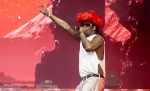 Rapper with bright red hair and sunglasses performs on stage in a white tank top and sparkly pants.