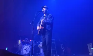 A man in a blue denim jacket and baseball cap sings and plays an acoustic guitar on a stage lit with blue light.