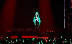 A digital anime character with long teal hair performs on a stage lit by red and green lights, with an audience holding glow sticks.