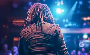 A person with dreadlocks stands facing away from the camera, illuminated by blue stage lights.