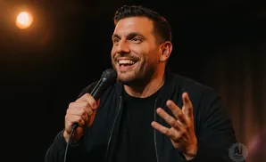 A man with a beard and short hair laughs while holding a microphone and gesturing with his hands.