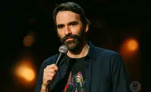 A man with a beard holds a microphone and wears a dark shirt with a colorful graphic.