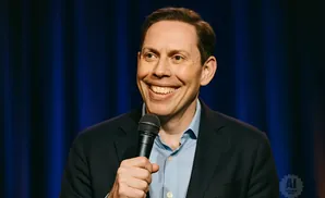 A man in a suit and blue shirt smiles widely while holding a microphone.