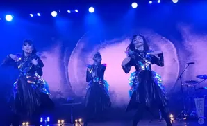 Three women in sparkly, futuristic outfits perform on a stage with blue lighting.