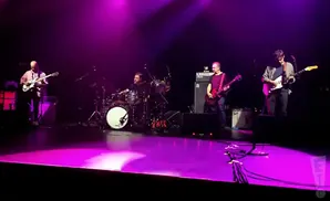 A band performs on a stage lit with purple lights. Four musicians are visible playing guitars and drums.