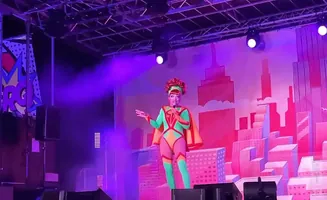 A drag queen in a green and orange superhero costume with a red wig performs on stage in front of a pink cityscape backdrop.