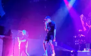 A male singer in a blue cap performs on stage with a guitarist to his left, under bright purple and blue lights.
