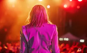 A woman with pinkish-blonde hair stands with her back to the camera, wearing a pink blazer on a brightly lit stage.