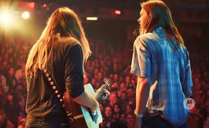Two men with long hair perform with guitars on stage to a cheering crowd.