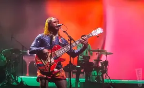 A guitarist with yellow dreadlocks plays a red guitar on a stage with a pink backdrop.
