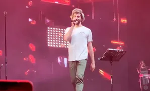 A man in a furry hat sings into a microphone on a stage with pink lights.