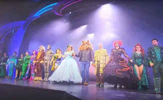 A cast in colorful costumes takes a bow on stage, with smoke and bright lights in the background.