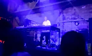 A DJ wearing headphones and a white shirt performs on a synthesizer in a dark, blue-lit venue.