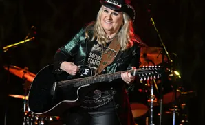 A blonde woman in a sparkly green jacket and cowboy hat plays a black 12-string guitar on stage.