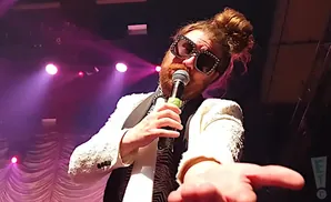 Man with messy bun, large bedazzled sunglasses, and a white jacket sings into a microphone on stage.