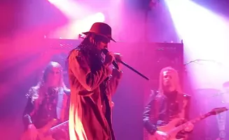 A band performs on stage with pink and purple lighting, a lead singer in a hat and long coat holds a microphone.