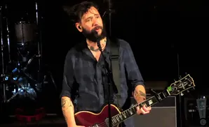 A man with a beard and tattoos plays a red guitar and sings into a microphone on a dark stage.
