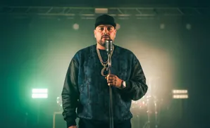 Man in black cap and patterned sweatshirt holds a microphone, wearing two gold chains.